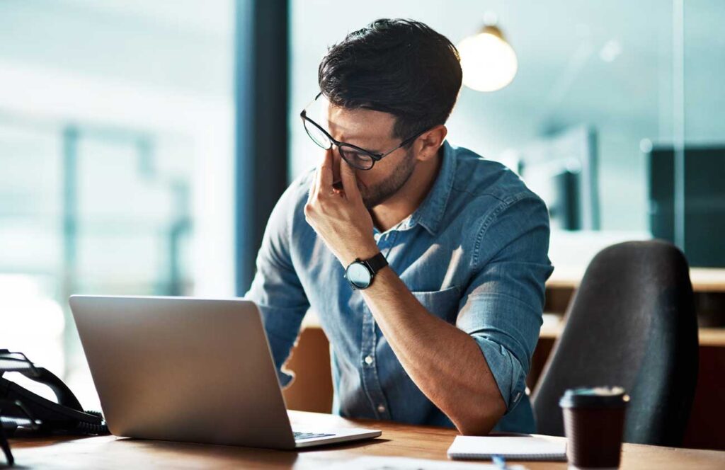 A stressed man rubbing his eyes while working on a laptop.
