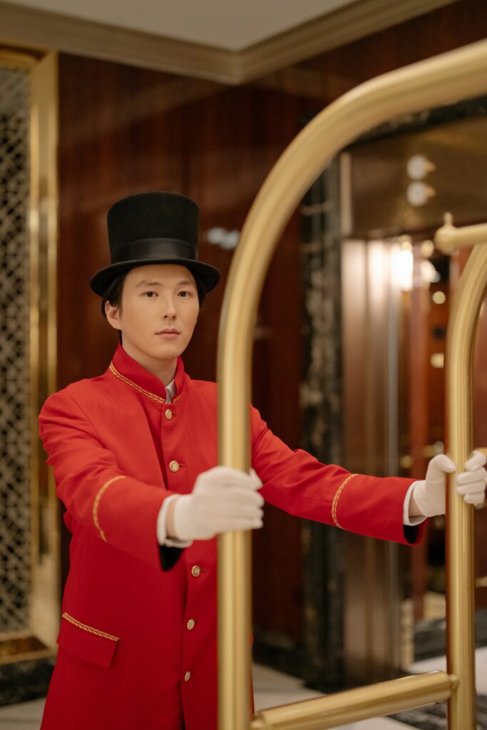 A hotel bellhop in a red uniform and top hat stands confidently.